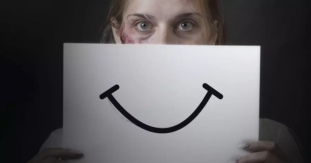 Foto de uma mulher loira, com olhos claros e hematomas aparentes no rosto, segurando um cartaz com o desenho de um sorriso mostrando como as mulheres disfarçam quando sofrem violência doméstica