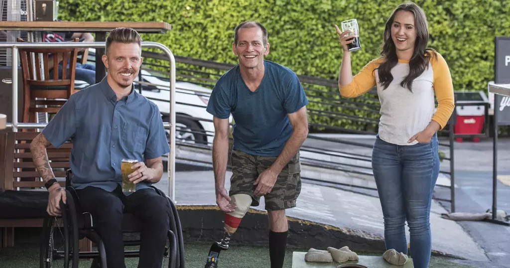 Um homem em uma cadeira de rodas, um segundo homem em pé e com uma prótese na perna direita e uma mulher em pé segurando um copo de refrigerante, como se estivessem em um happy hour da empresa mostrando a importância da inclusão de todas as pessoas