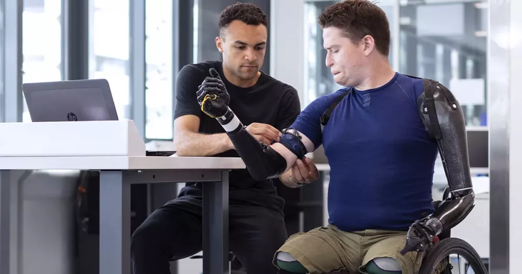 Dois homens em um escritório onde um deles ajusta as próteses de braço do outro, mostrando a luta das pessoas com deficiência