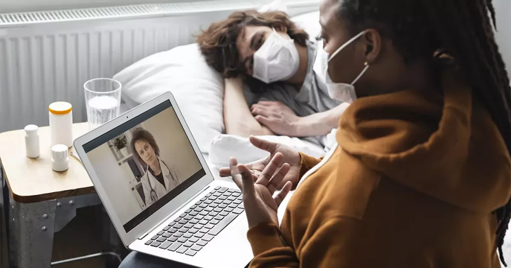 Foto de uma mulher negra, utilizando máscara e falando com uma médica através de um notebook em uma consulta por telemedicina. Ao fundo, um rapaz branco, também com máscara, deitado em uma cama e aparentando estar doente.