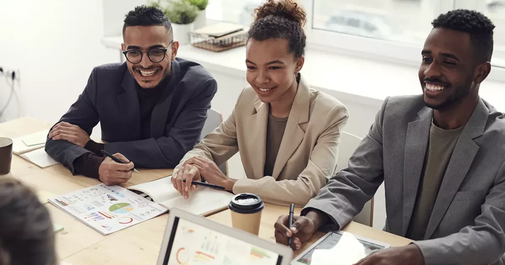Foto de três colaboradores felizes, sendo dois homens e uma mulher no meio, sentados a mesa do escritório e sorrindo. A foto representa como uma boa gestão de pessoas é importante para os negócios.