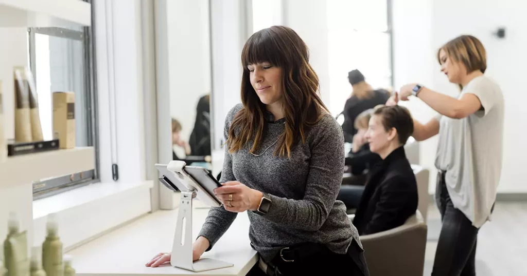 Foto de uma mulher mexendo em um tablet em destaque e ao fundo cabelereiras e clientes em um salão de beleza. A foto representa o fato de que até pequenas empresas podem contratar um plano de saúde empresarial.