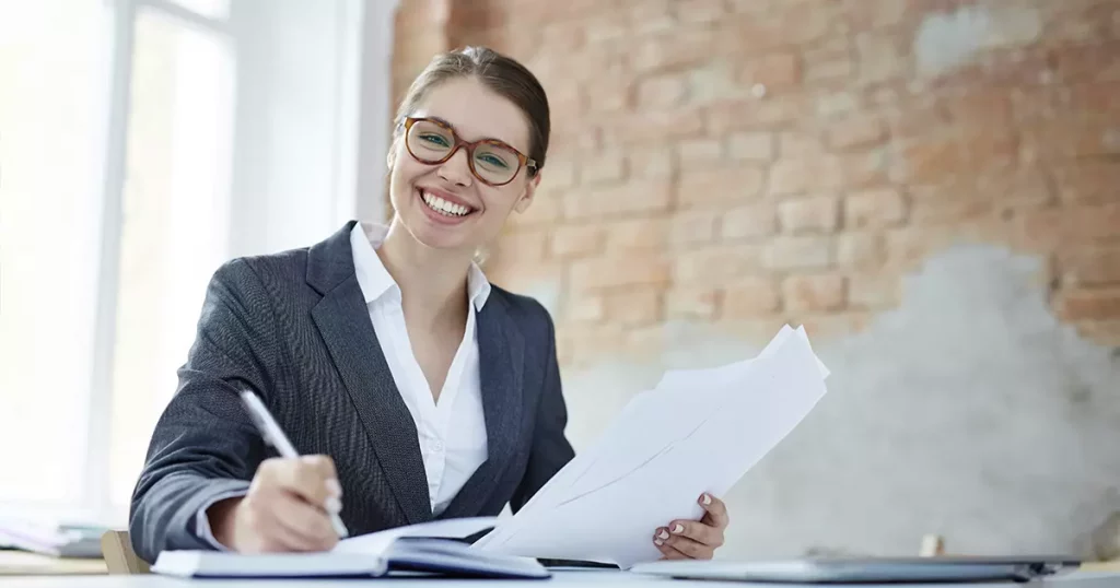 Foto de uma mulher branca, com cabelo preso, usando óculos arredondados e um blazer cinza escuro por cima de uma camisa social branca, sorrindo e fazendo anotações em papeis representando o mapeamento de processos contábeis.