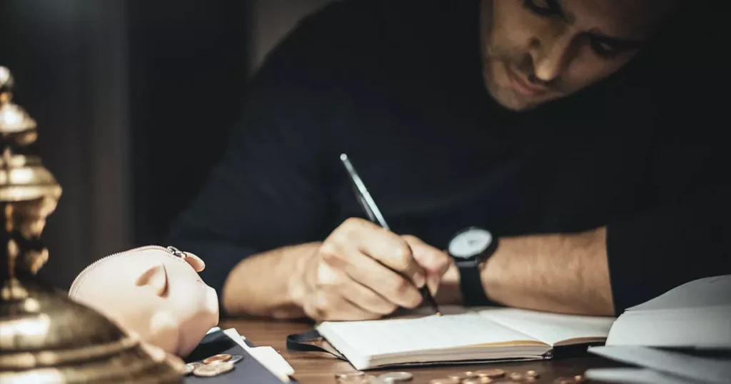 Foto de um homem anotando cálculos em um caderno com um cofre de porquinho do lado mostrando como o controle de custos pode ajudar na redução de custos.