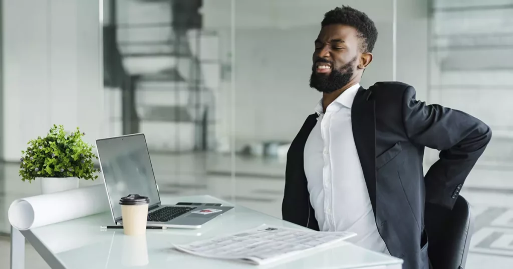 Foto de um homem negro, usando terno e sentado em sua cadeira de trabalho aparentando sentir dor nas costas por falhas de ergonomia no trabalho.