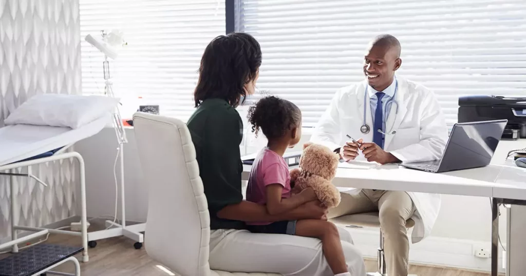 Foto de um consultório médico com um médico negro sentado em sua mesa e na frente da mesa, mas de costas para a foto, uma mulher segurando uma criança no colo que segura um ursinho de pelúcia em suas mãos. A imagem serve para ilustrar as diferenças entre plano e seguro saúde.