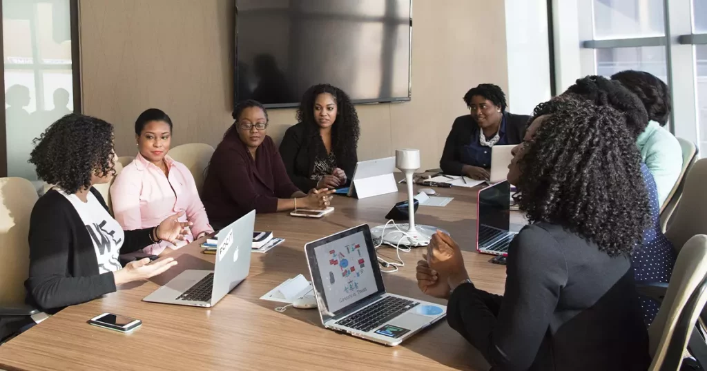 Foto de oito mulheres negras sentadas em volta de uma mesa de reunião em uma empresa mostrando a importância da liderança feminina.