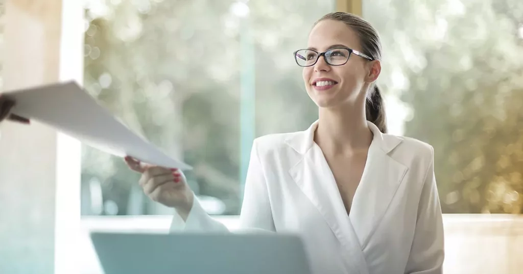 Foto de uma mulher branca, de óculos, como cabelo amarrado em um rabo de cavalo e usando um blazer branco, sentada e recebendo alguns papeis de uma outra pessoa que não aparece na imagem. Ela representa uma funcionária realizando a gestão da saúde de uma empresa.