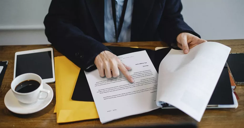 Foto das mãos de um homem sobre um contrato em cima de uma mesa de escritório, como se estivesse acompanhado a leitura com os dedos. Em volta do contrato existe um tablet, uma xícara de café, um celular e algumas pastas. A imagem representa as novas regras de portabilidade de carência determinadas pela ANS.