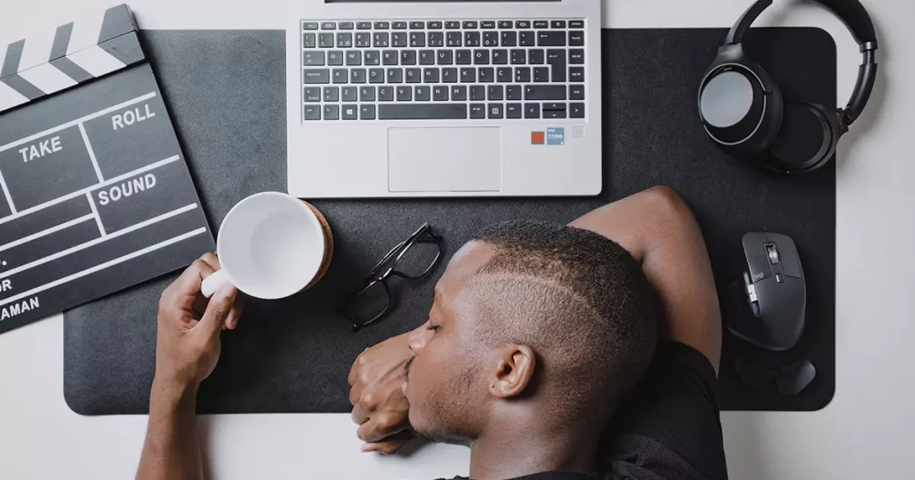 Foto de um rapaz negro dormindo na mesa de trabalho, segurando uma caneca vazia e com um notebook a sua frente representando como a saúde mental no trabalho impacta na produtividade dos funcionários,