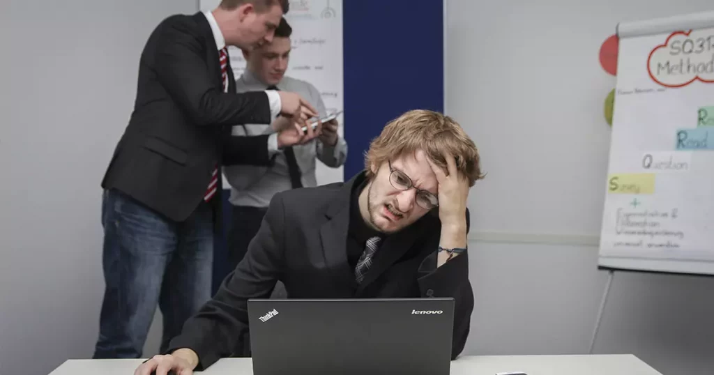 Foto de um homem loiro, usando óculos e terno preto olhando para um notebook cm uma cara de estresse enquanto coloca a mão esquerda na cabeça, representando uma das causas da síndrome de burnout. Ao fundo e do lado esquerdo da imagem, mais dois homens em pé olhando um tablet e do lado direito um flipchart com anotações.