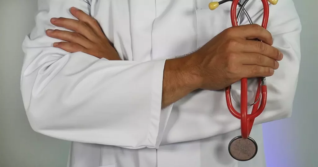 Foto dos braços cruzados de um médico segurando um estetoscópio vermelho.