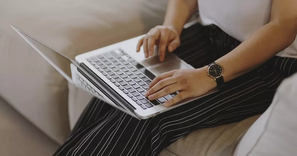 Foto do tronco, pernas e braços de uma mulher sentada em um sofá com um notebook no colo pesquisando sobre SulAmérica ou Bradesco.