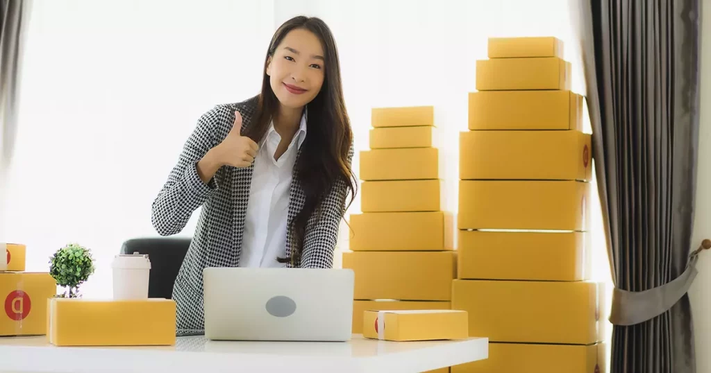 Foto de uma mulher oriental com algumas caixas atrás dela e um notebook a sua frente mostrando a importância do controle de estoque.