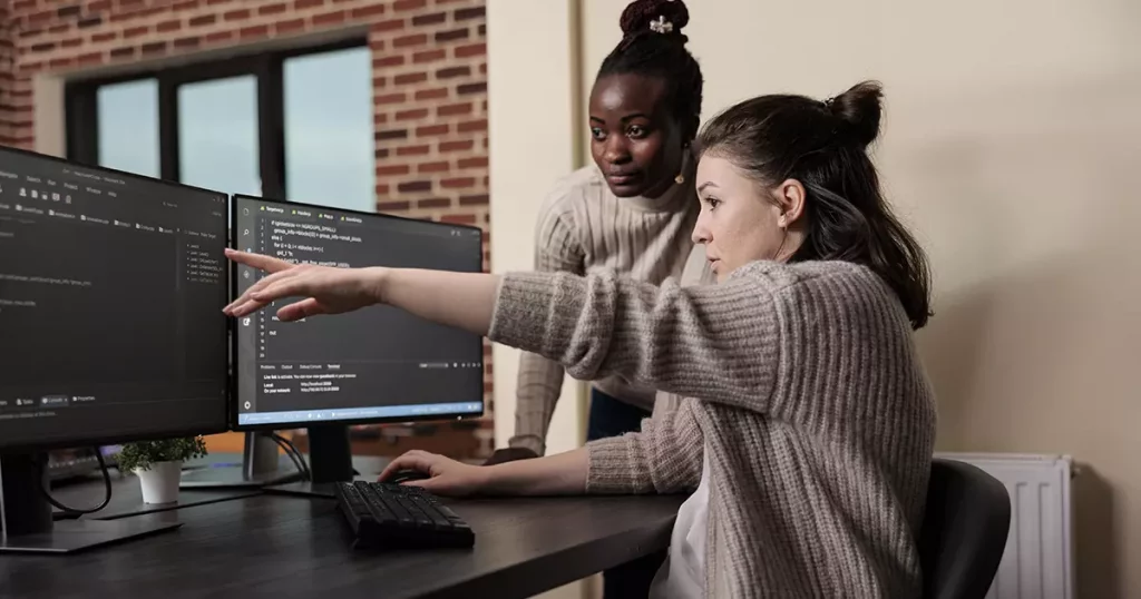 Duas mulheres, uma negra e uma branca, trabalhando juntas em um computador com duas telas e com códigos aparecendo nelas, representando o fato de que qualquer tipo de empresa podo contratar um seguro cibernético. 
