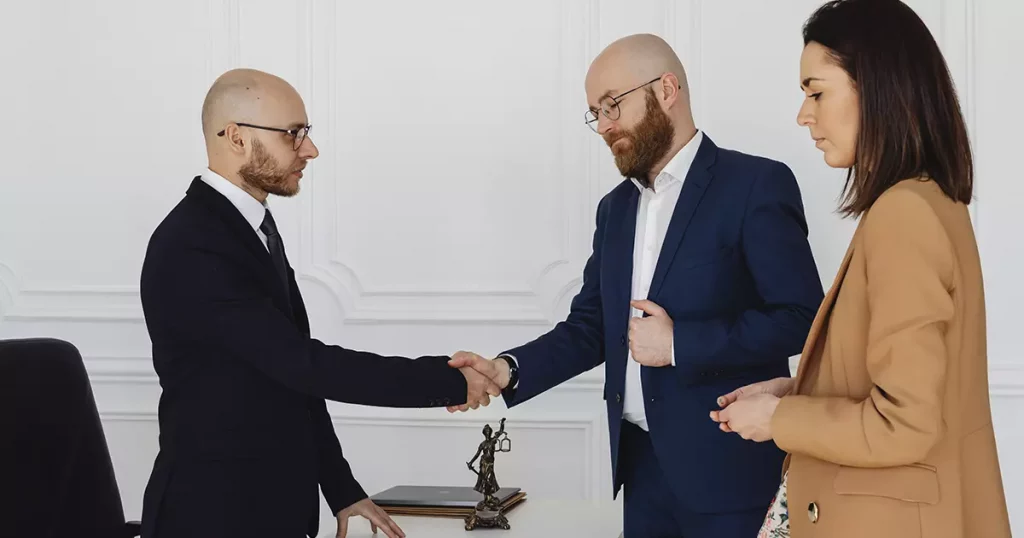 Dois homens brancos e carecas, vestindo ternos e óculos, apertando as mãos como se estivesse fechando um negócio. Ao lado do homem da direita está uma mulher branca, cabelo liso na altura do ombro e um blazer bege olha a negociação dos dois homens.