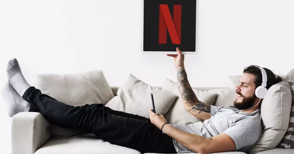Homem branco deitado em um sofá, usando um headphone enquanto olha para o celular e segurando uma placa com a letra "N" do logotipo do Netflix
