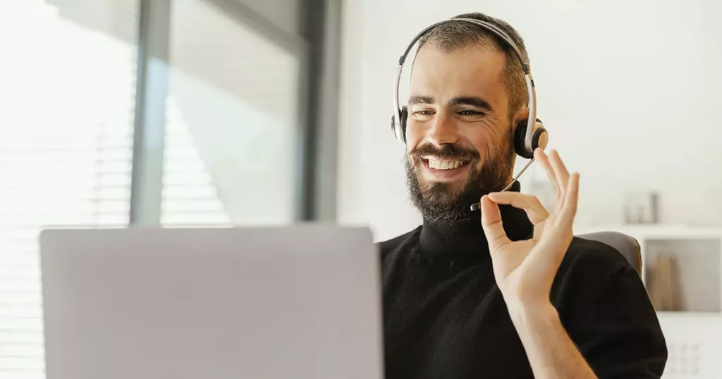 Homem branco, com barba, cabelo curto, usando um headset e blusa preta, fazendo sinal de OK para o notebook durante reuniões online.