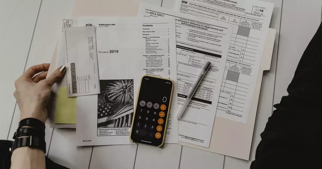 Foto da mão esquerda de uma pessoa segurando algumas contas e um celular com a calculadora aberta representando o controle de gastos para recuperar a saúde financeira.