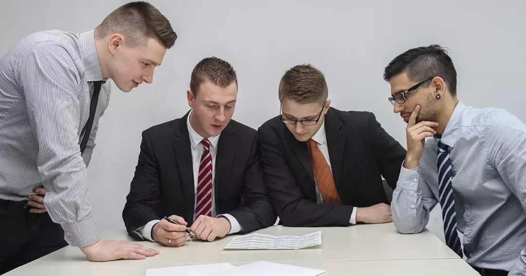 Foto de quatro homens de negócios, sendo dois vestindo terno e gravata e dois apenas com camisa social de manga longa e gravatas. Todos sentados em volta de uma mesa olhando para papéis, estudando sobre os tipos de previdência privada para a empresa deles.