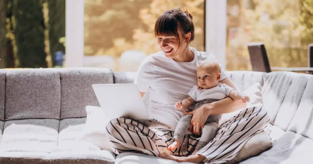 Mulher com cabelo castanho e franja, sentada no sofá, com o notebook apoiado em uma perna e um bebê na outra representando o funcionário trabalhando no home office