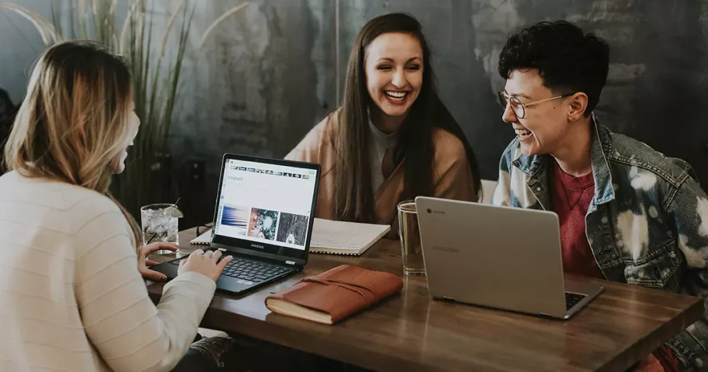 Fotografia de três mulheres trabalhando juntas em uma mesa sorrindo. Uma delas é loira e está de costas para a câmera mexendo em um notebook aberto. Outra é branca, cabelo escuro e longo e está anotando algo em um caderno. A terceira é branca, cabelo bem curto, usa óculos e uma jaqueta jeans por cima de uma camiseta vermelha e está de frente para um notebook aberto também.