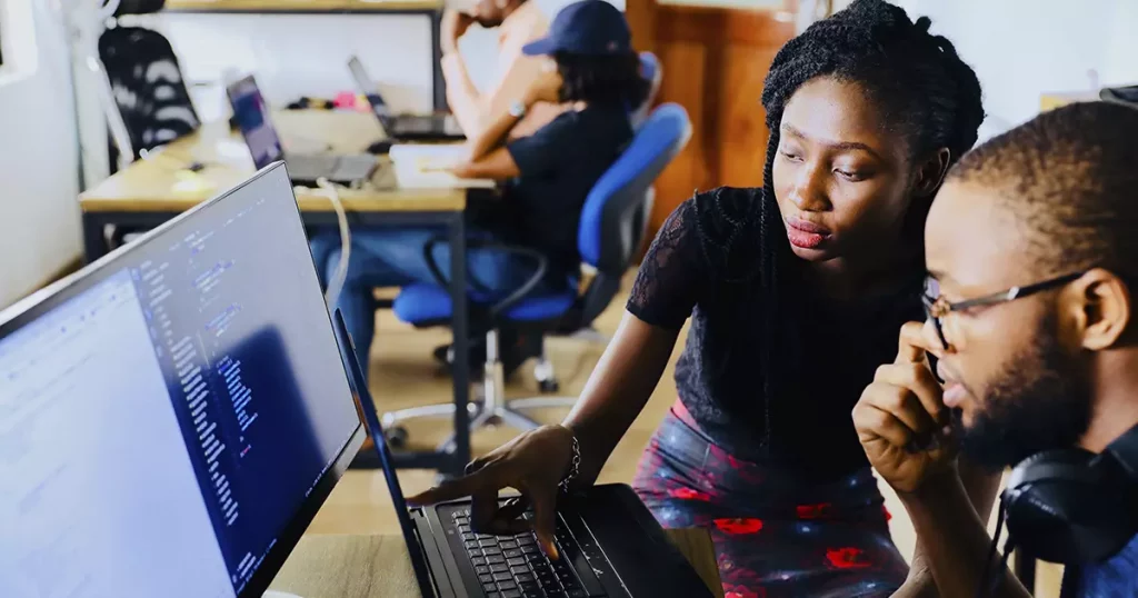 Um homem e uma mulher, ambos negros, olhando para a tela de um computador e um notebook com códigos, representando diversas pequenas empresas