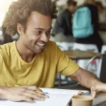 Fotografia de um homem negro sentado numa mesa de escritório mexendo num notebook enquanto anota algo em seu caderno. O homem está sorrindo e vestido com uma camiseta amarela.
