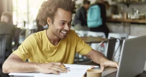 Fotografia de um homem negro sentado numa mesa de escritório mexendo num notebook enquanto anota algo em seu caderno. O homem está sorrindo e vestido com uma camiseta amarela.