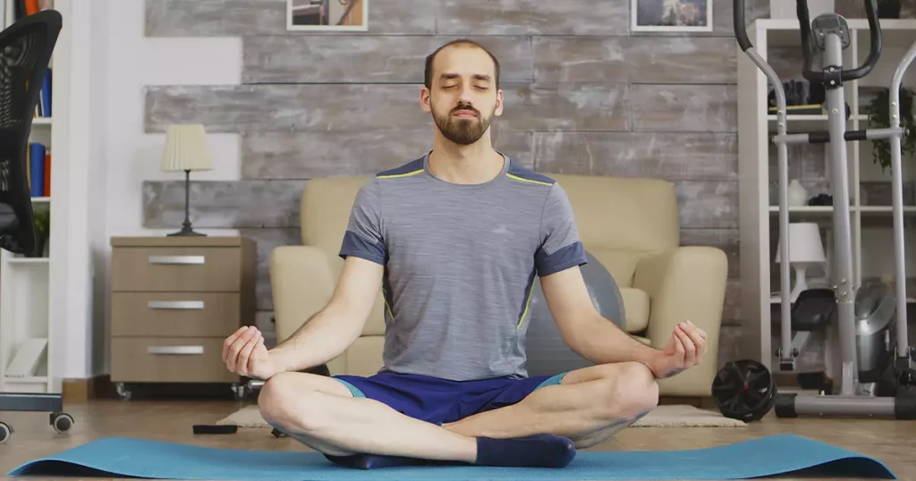 Homem branco vestido com roupas de academia em tons de azul sentado em pose de yoga: pernas cruzadas, olhos fechados e mãos apoiadas nos joelhos.
O homem está sentado sobre um tapete de yoga num quarto.